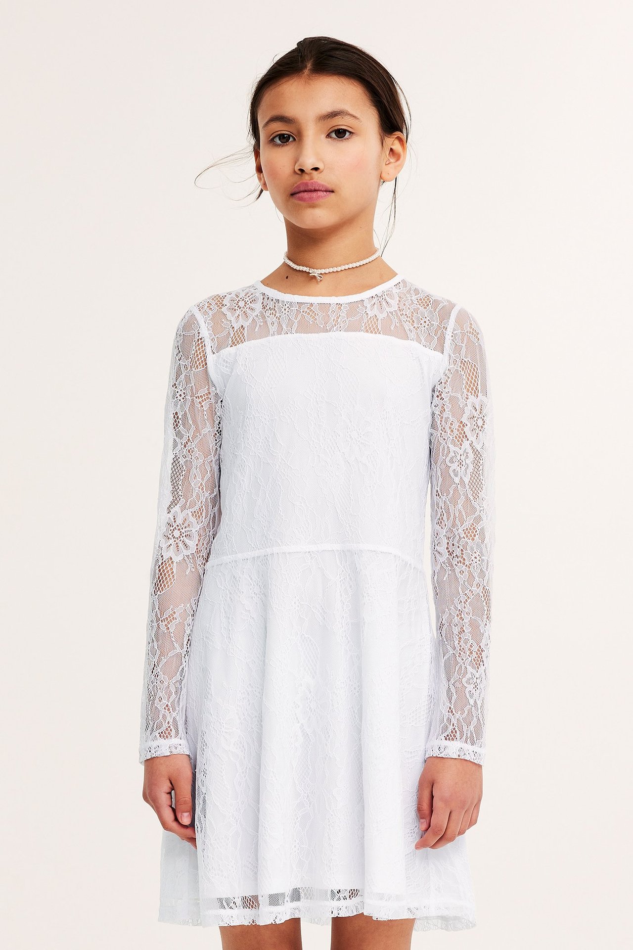 Frontvisning av et barn i en hvit langermet blondekjole og et perlekjede.