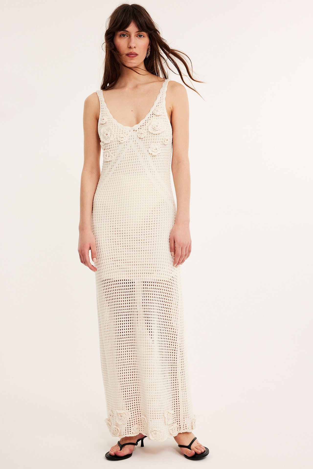 Frontvisning av en kvinne i lang beige heklet maxikjole med påsydde blomster og svarte sandaler.
