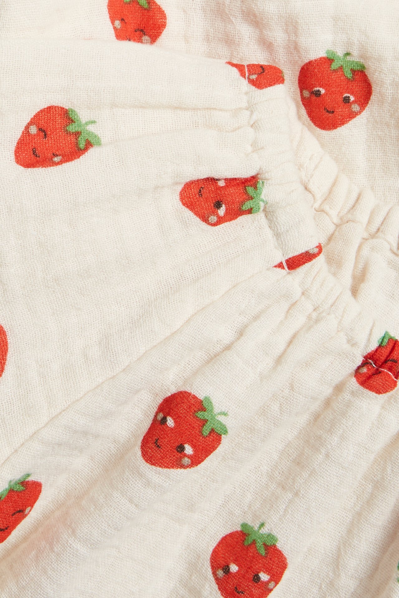 Närbild på en babyklänning i off-white muslin med rött jordgubbsmönster.