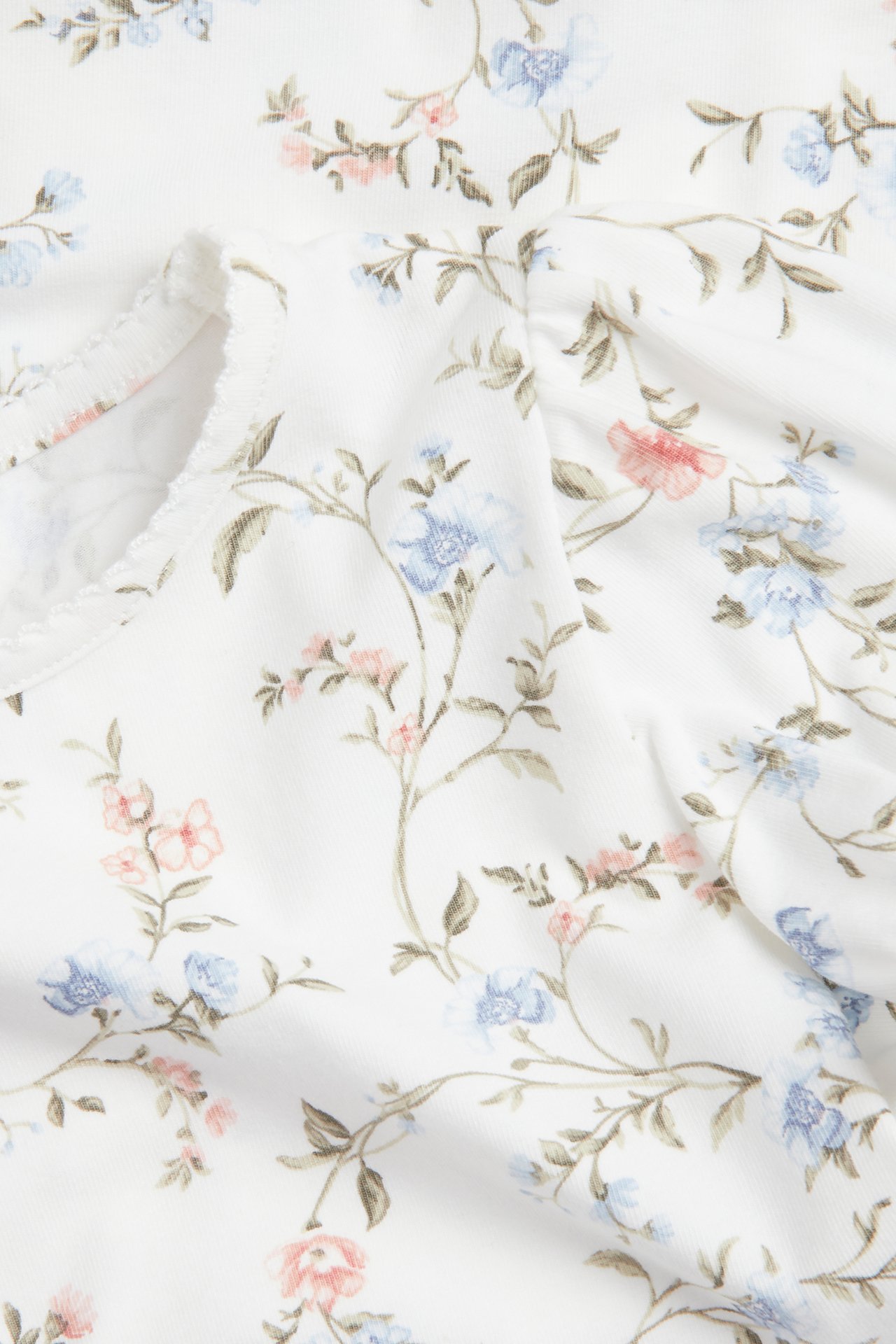 Nærbilde av en offwhite kortermet blomstret topp med blå/rosa blomster, puffermer og picotkant.