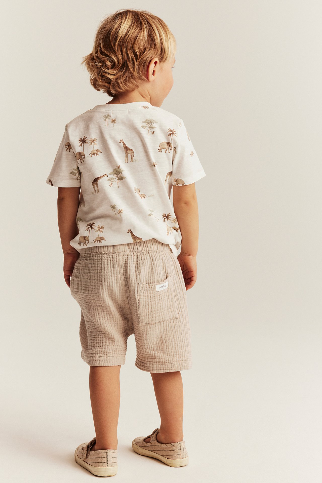 Bakifrån: ett barn i beiga vävda shorts med resår, bakficka och en vit t-shirt med djurmönster.