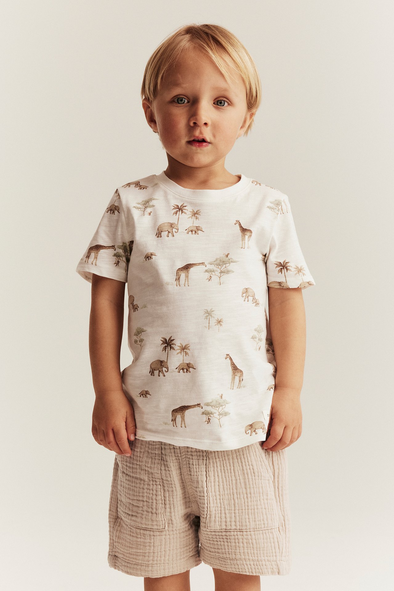 Edestäpäin, lapsi yllään beiget kuvioidut kudotut shortsit ja valkoinen eläinkuvioinen t-paita.