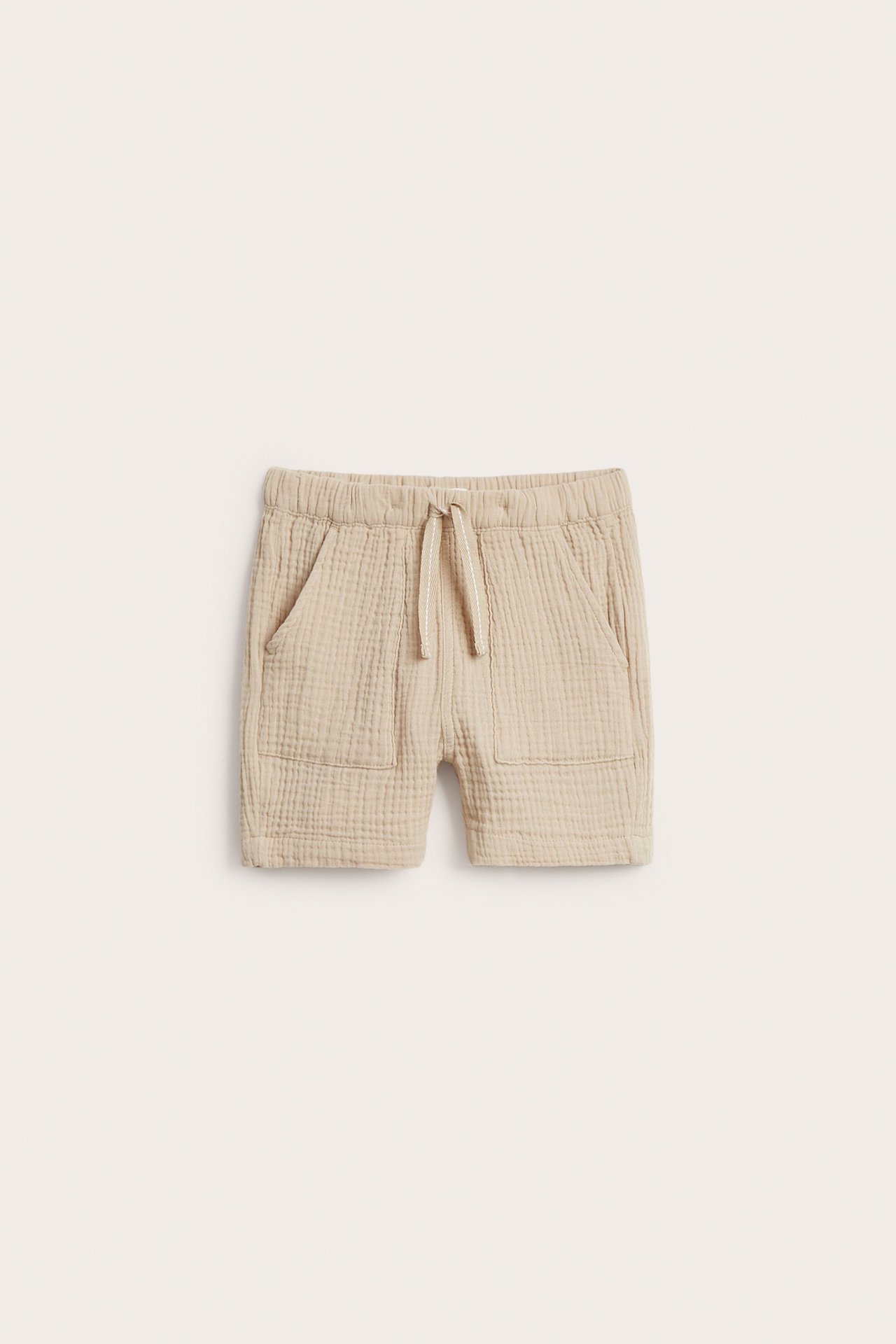 Framifrån: beiga vävda bomullsshorts för barn med resår i midjan, dragsko och påsydda framfickor.