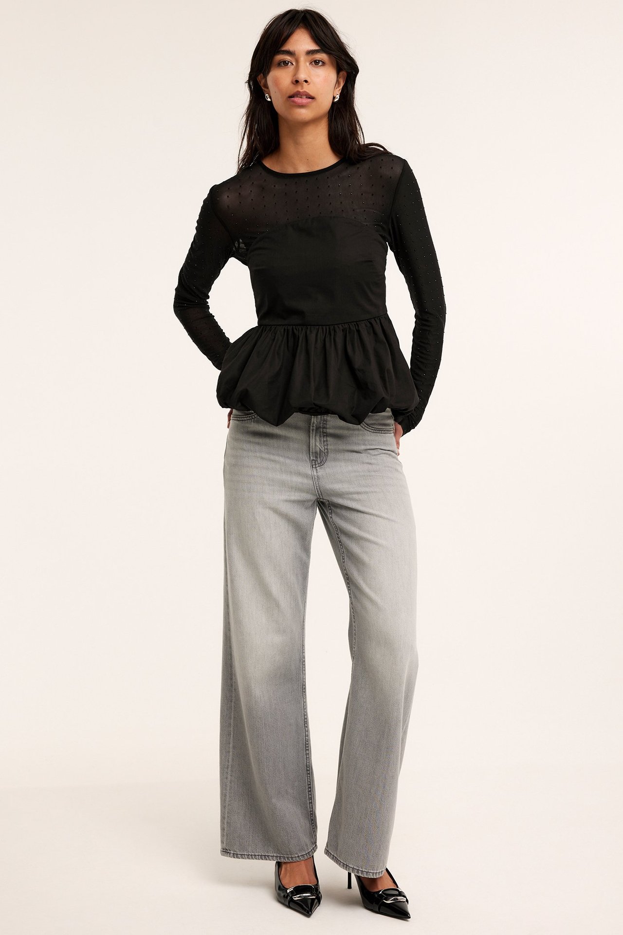 Frontbilde: kvinne i svart peplumtopp med prikkete ermer, grå vide jeans og svarte hæler.