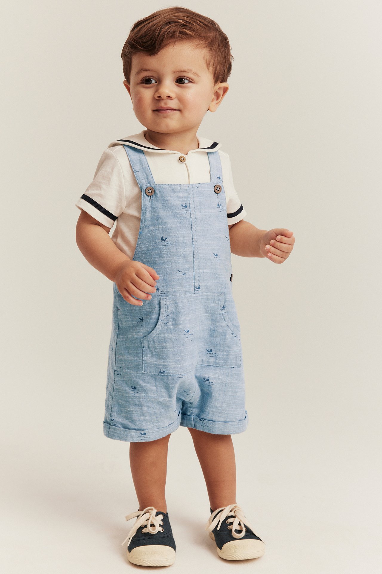 Frontvy av baby i ljusblå mönstrade chambray hängselshorts över vit sjömanskrage-topp och mörkblå sneakers.