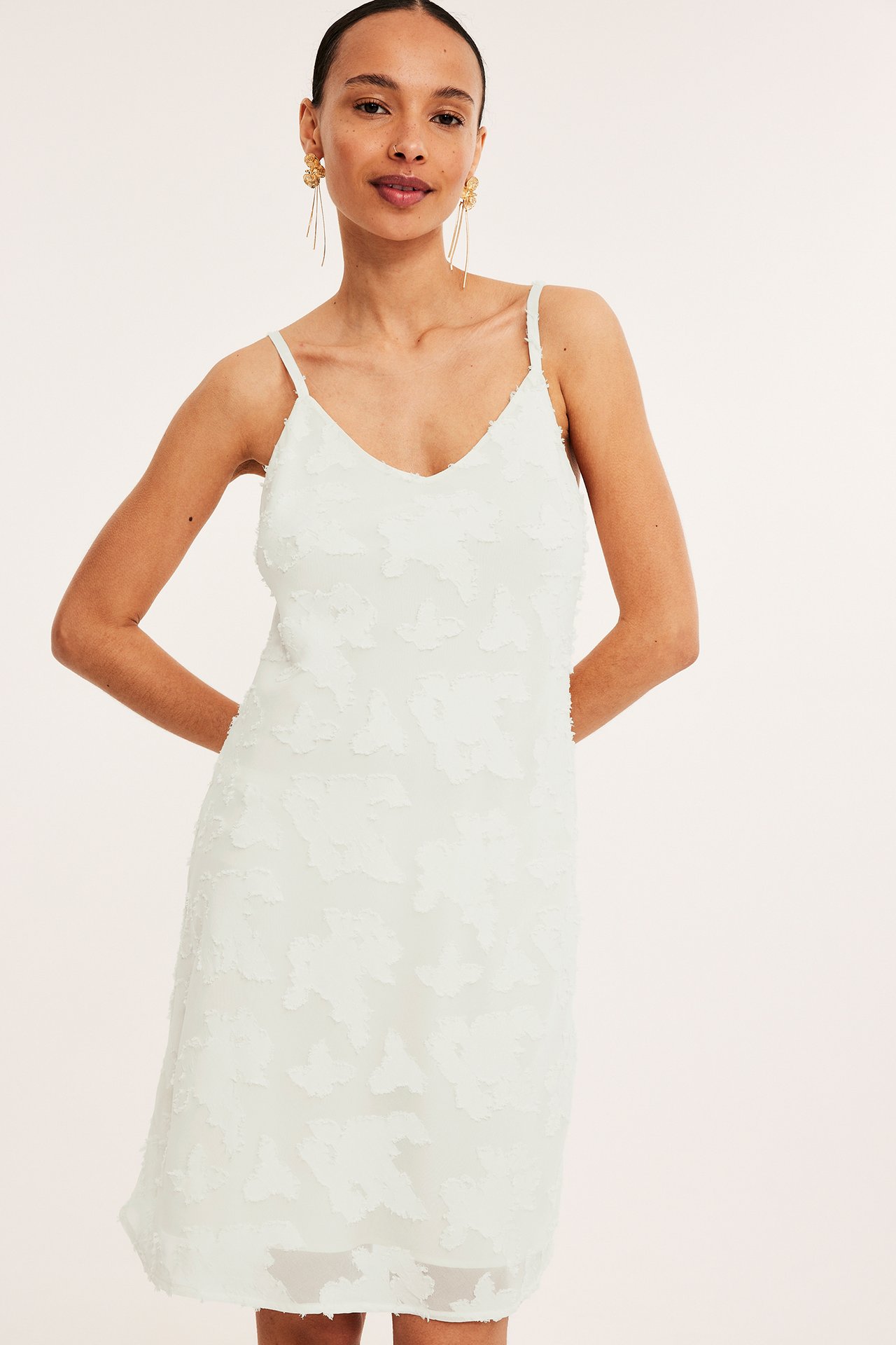 Framifrån: kvinna i kort offwhite jacquardmönstrad slipklänning och långa guldfärgade örhängen.