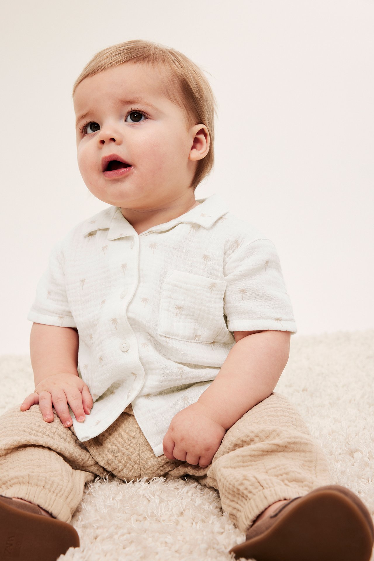 Framifrån: baby i offwhite muslin skjorta med palmprint, beiga muslinbyxor och bruna skor.