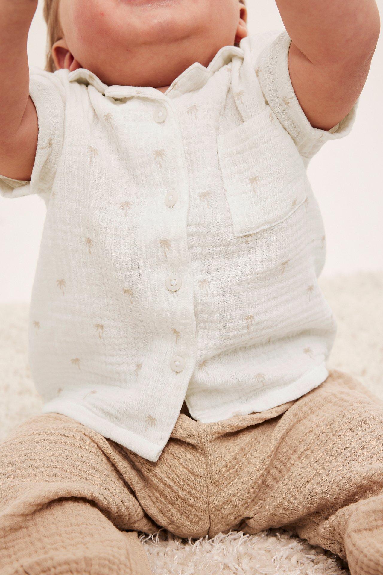 Närbild framifrån på baby i offwhite muslin skjorta med palmträdsmönster och ljusbruna muslinbyxor.