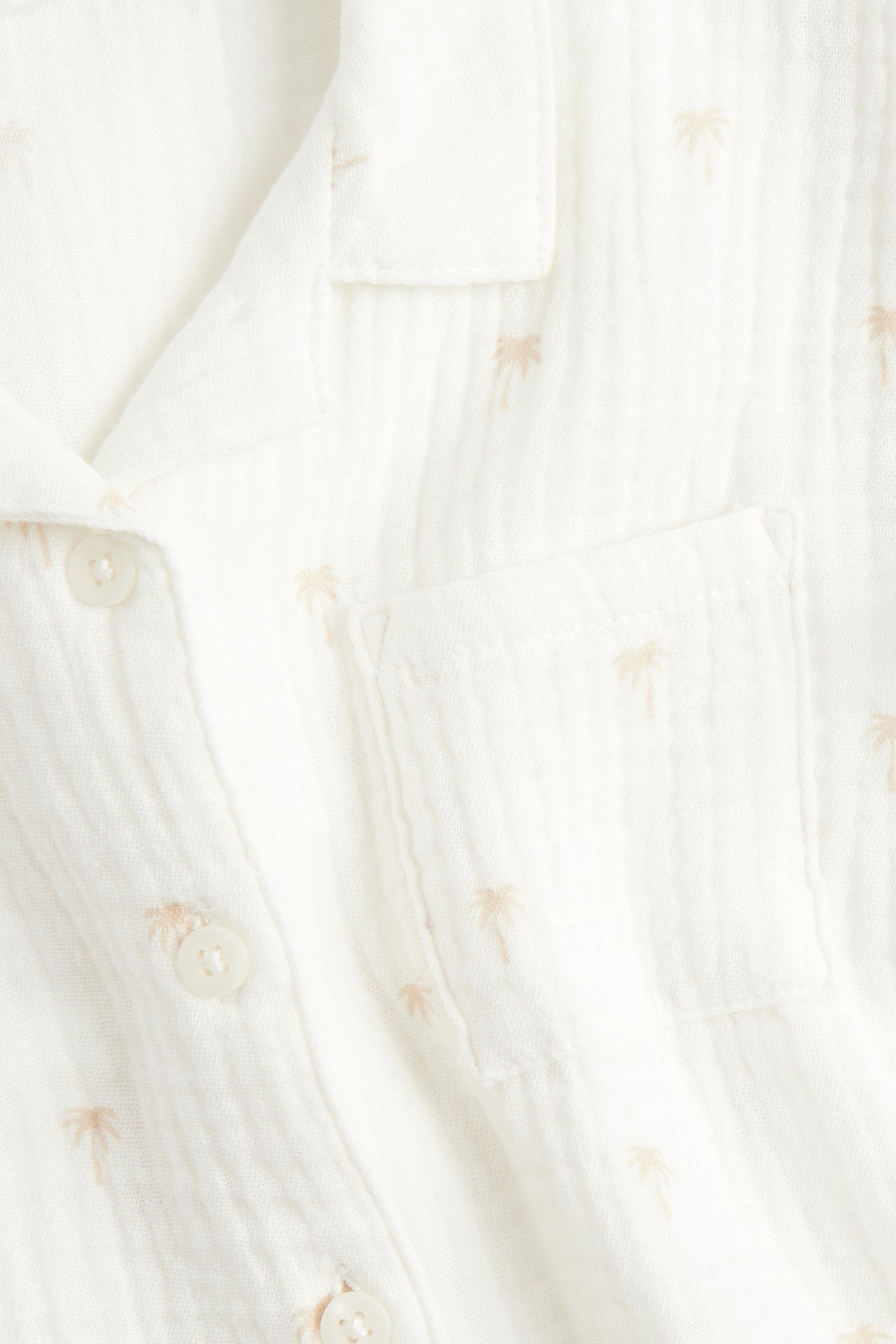 Närbild framifrån på en offwhite baby-muslinskjorta med ljusbrunt palmträdsmönster, bröstficka och knappar.
