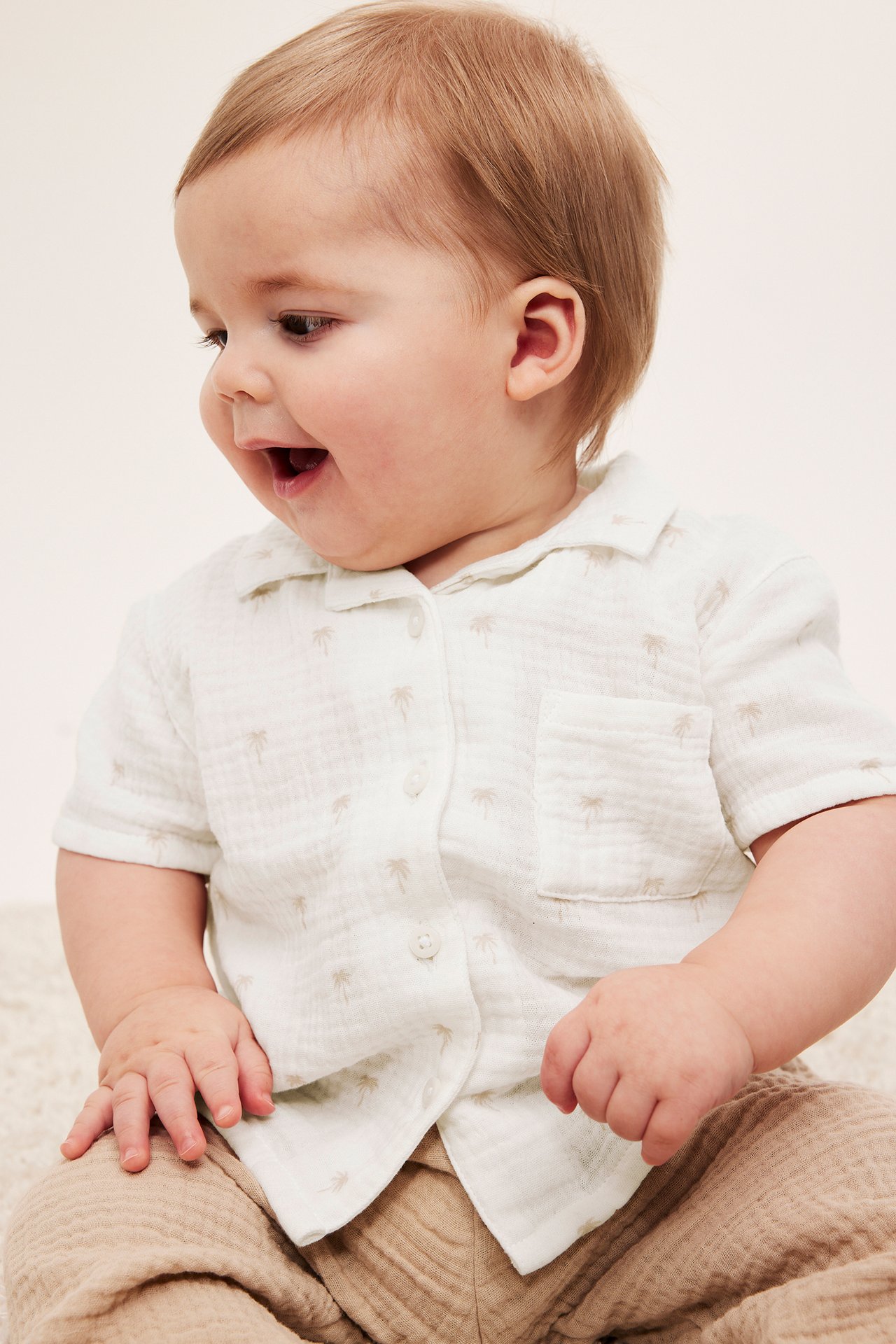 Frontvy av leende baby i offwhite muslin skjorta med palmprint och ljusbruna byxor.