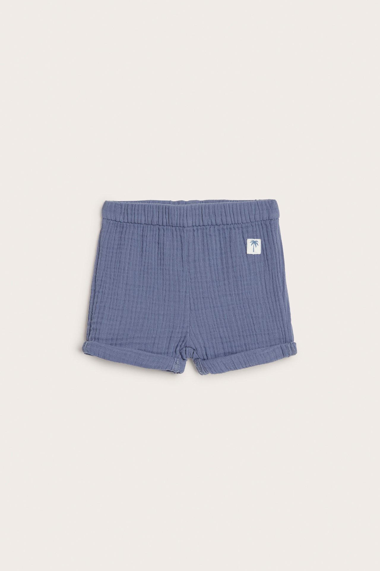 Frontvy av blå baby-muslinshorts med resår i midjan, uppvikta benslut och en palm-etikett.