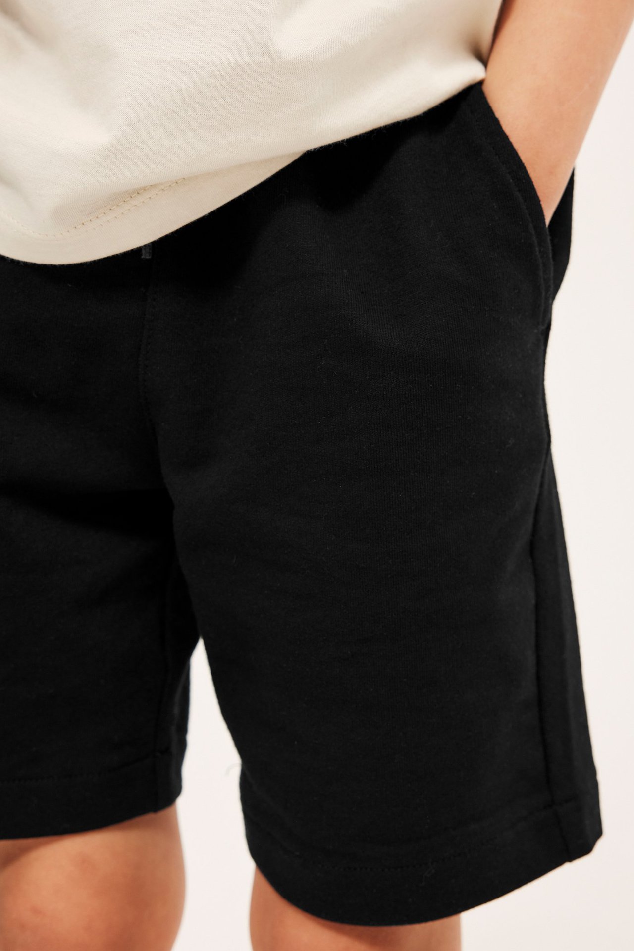 Frontvy av barn i enfärgade svarta bomullsshorts med hand i fickan och ljus t-shirt.