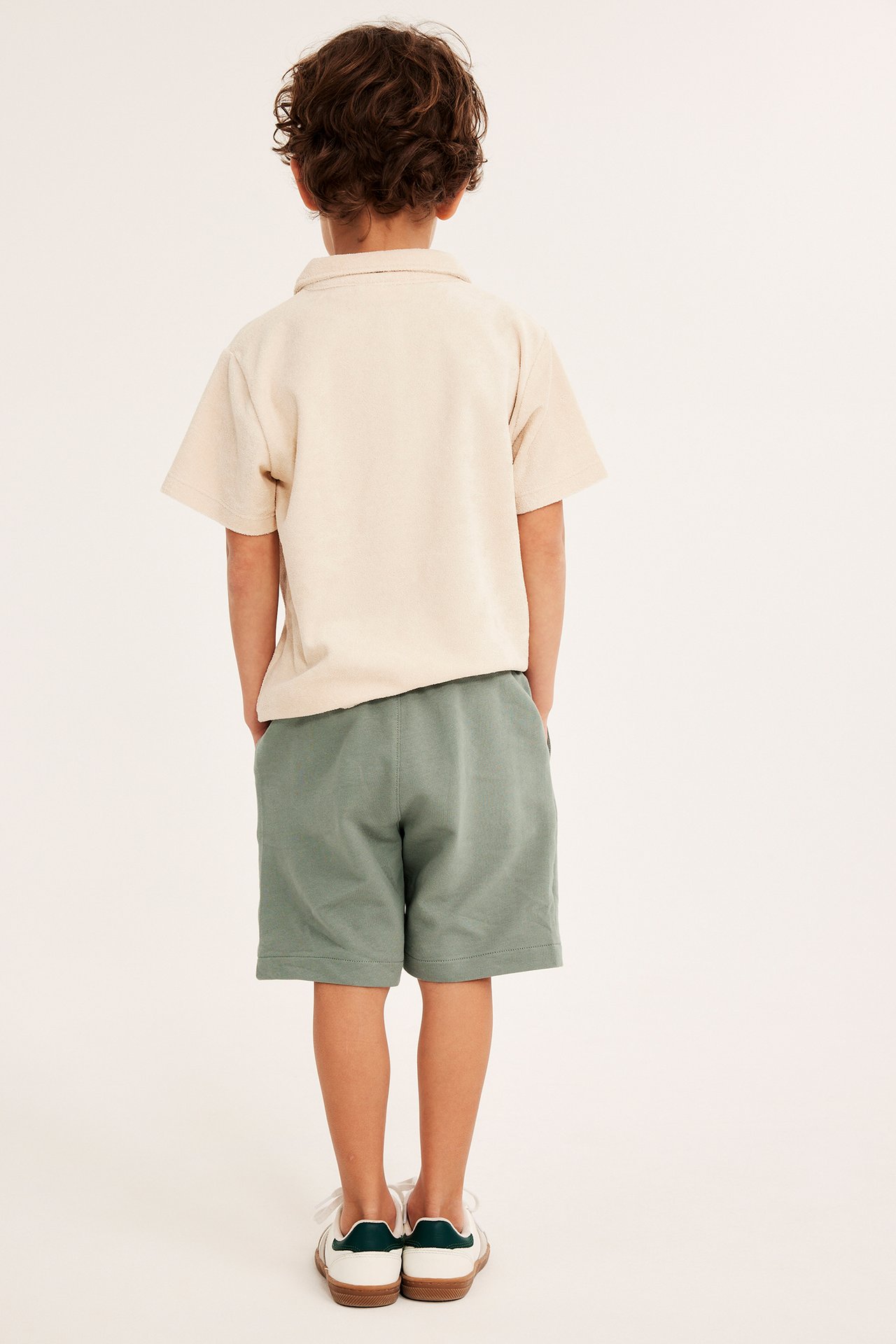 Bakifrån, ett barn i mörkgröna bomullsshorts, en ljusbeige pikétröja och vita sneakers.