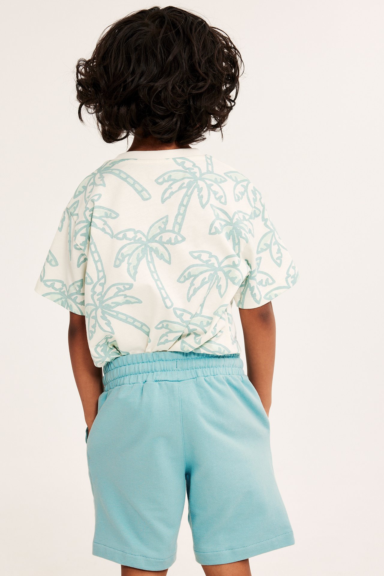 Bakifrån: ett barn i ljusgröna bomulls-mjukisshorts med händerna i fickorna och en vit t-shirt med palmtryck.