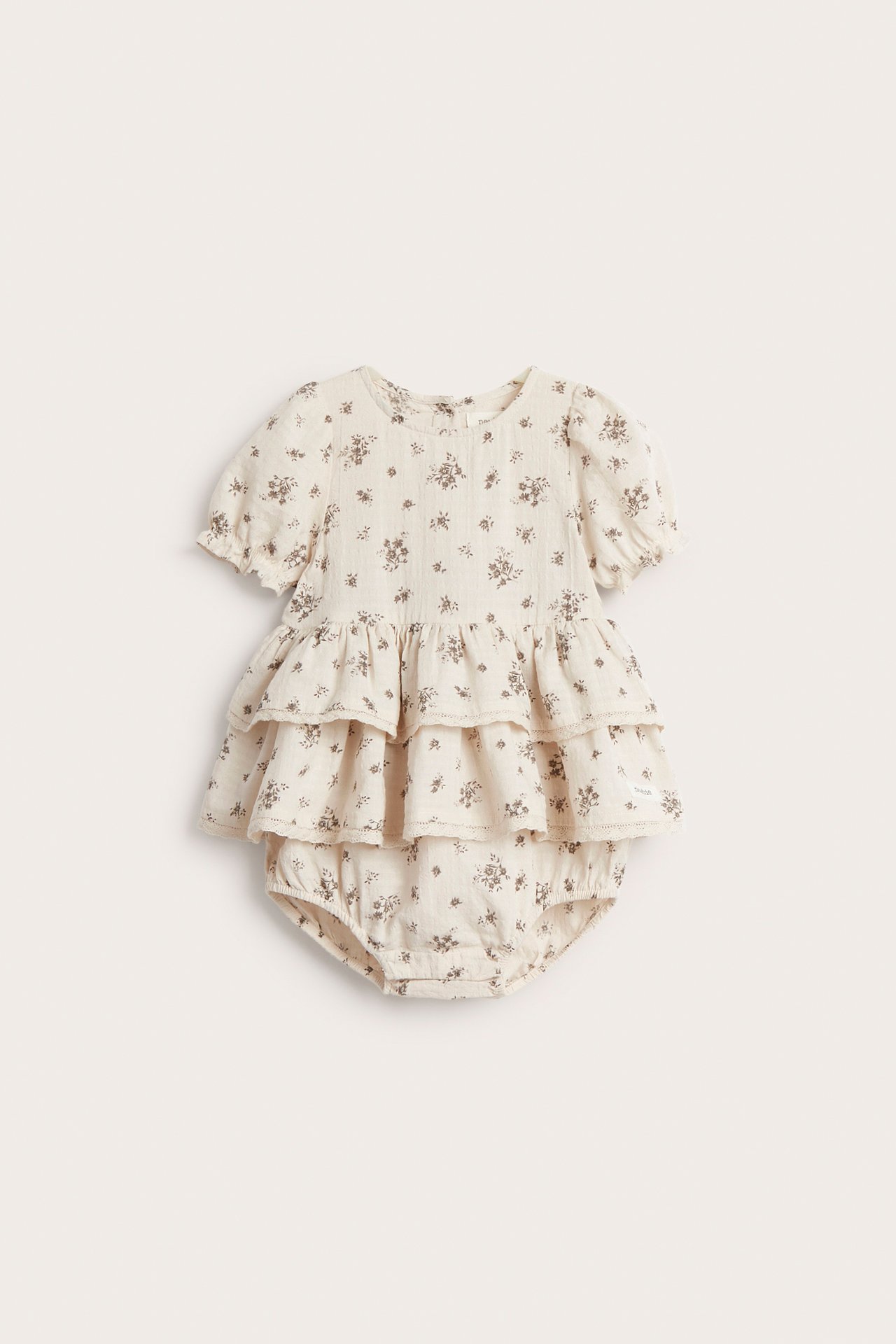 Edestä, vauvan vaaleanbeige kukkakuvioinen puuvillaplaysuit röyhelökerroksilla ja lyhyillä joustavilla hihoilla.