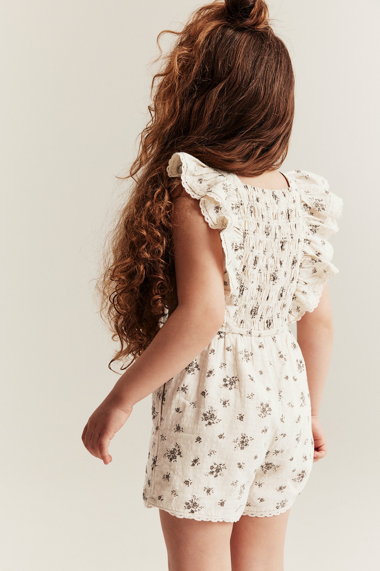 Bakifrån: ett barn i en ljusbeige blommig bomullsjumpsuit med smock, volangärmar och spetskant.