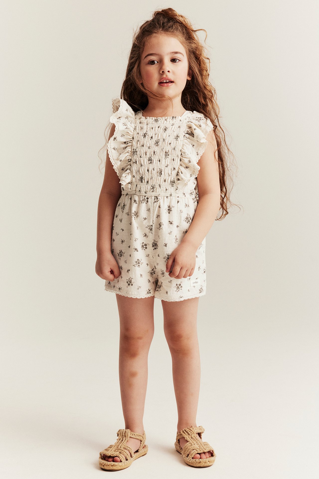 Framifrån: Barn i ljusbeige blommig kortärmad jumpsuit med smockad överdel, volangaxlar och vävda sandaler.