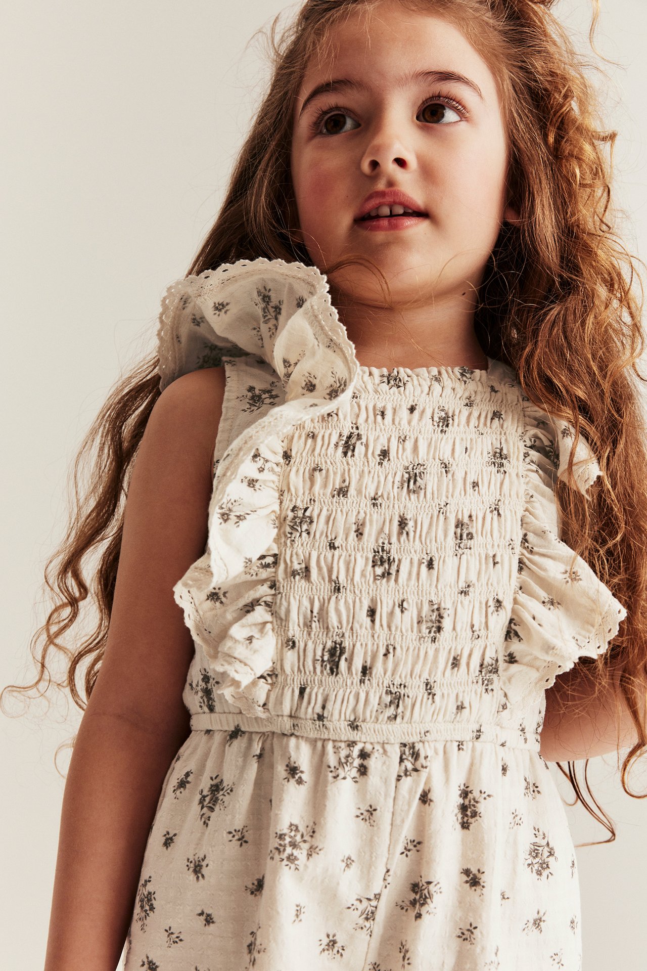 Närbild framifrån på ett barn i en ljusbeige blommig bomullsjumpsuit med smockad överdel och volangärmar med spets.
