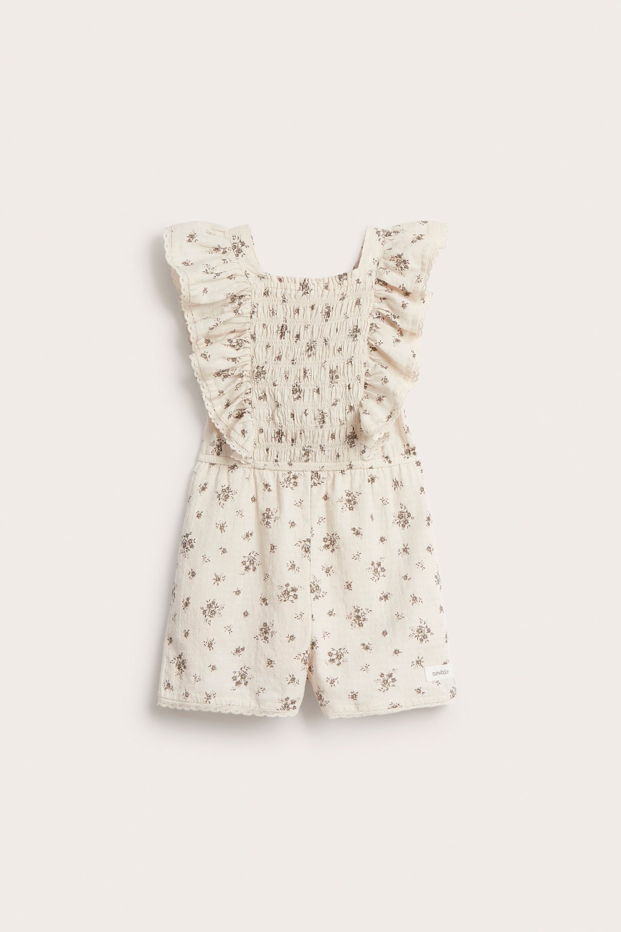 Frontvy: Barnens ljusbeige blommiga bomullsjumpsuit med smockad överdel, volangaxlar och spetskantade korta ben.