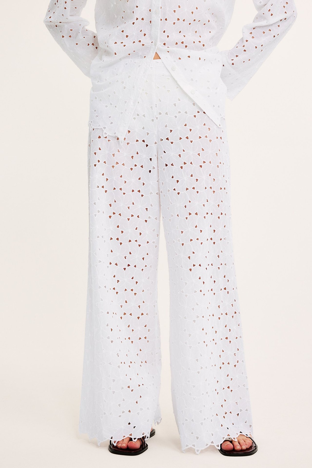 Edestä: Nainen yllään valkoiset leveät pitsihousut, yhteensopiva paita ja mustat sandaalit.