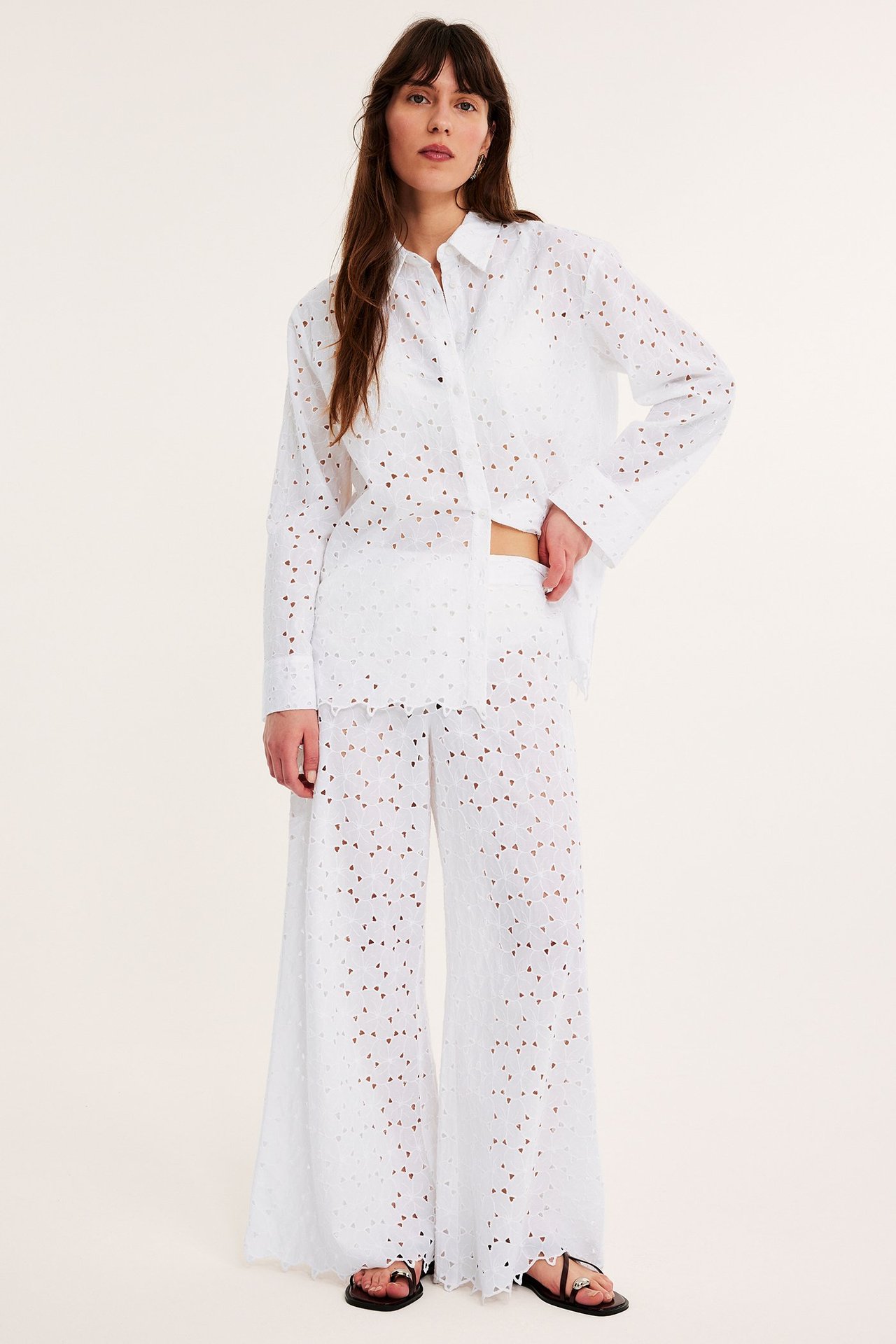 Naisen etunäkymä leveissä valkoisissa puuvillapitsihousuissa, yhteensopivassa paidassa ja mustissa sandaaleissa.