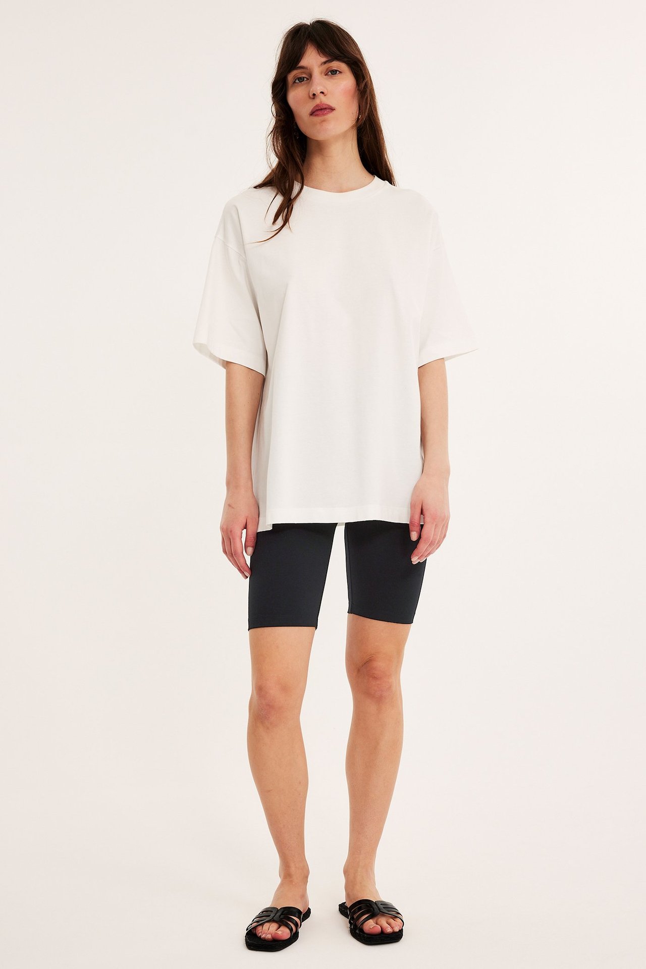 Forfra: Kvinne i svarte trikot-sykkelshorts, en hvit oversized t-skjorte og svarte sandaler.