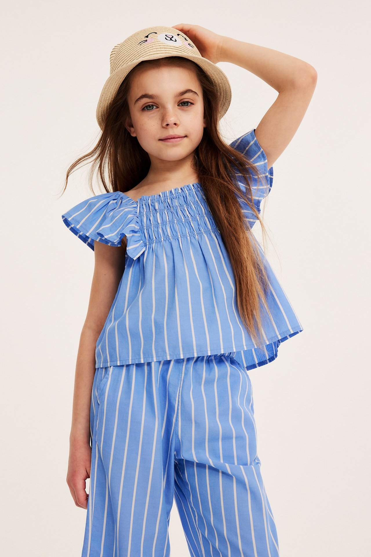 Widok z przodu dziecka w niebiesko-białej bluzce w paski z falbankami, pasujących spodniach i słomkowym kapeluszu.