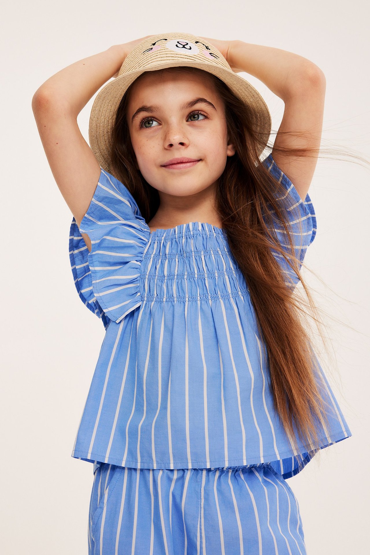 Widok z przodu dziecka w niebiesko-białym bawełnianym topie w paski z falbankami, pasujących szortach i słomkowym kapeluszu.