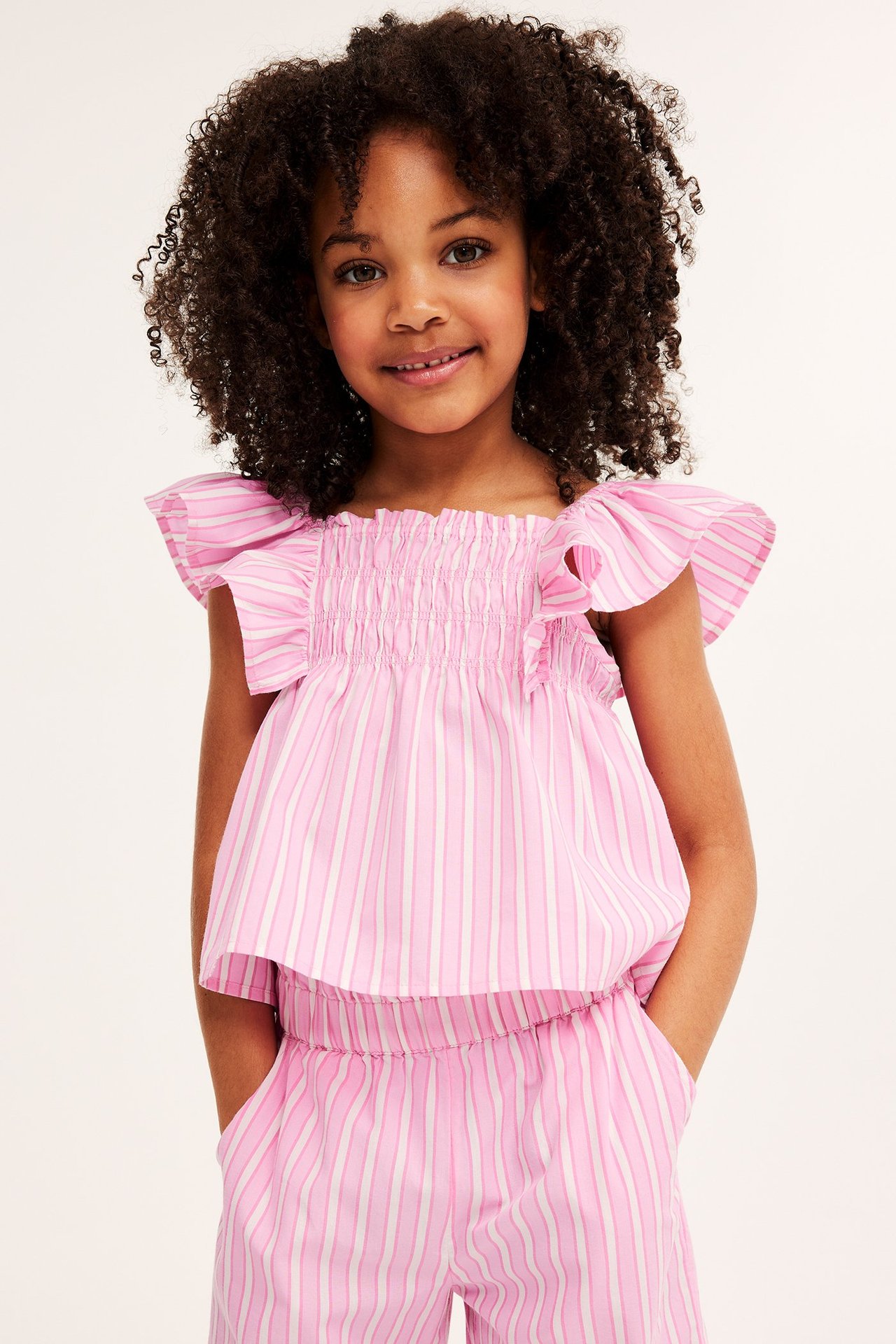 Widok z przodu dziecka w różowo-białej bluzce w paski z falbankami, marszczonym gorsetem i pasującymi spodniami.