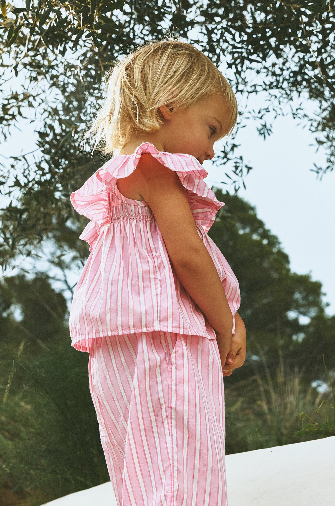 Widok z boku dziecka w różowo-białej bluzce w paski z falbankami i pasujących spodniach.