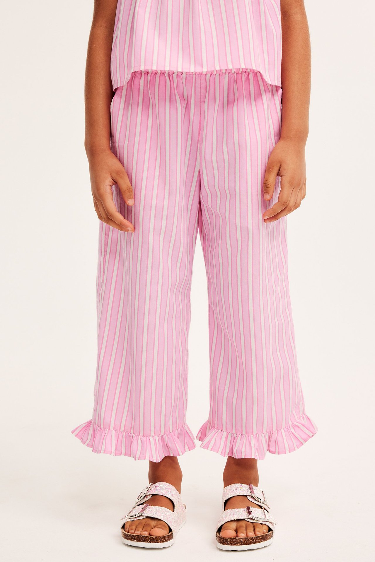 Widok z przodu: dziecko w różowo-białych bawełnianych culotte w paski z falbankami i różowych sandałach.