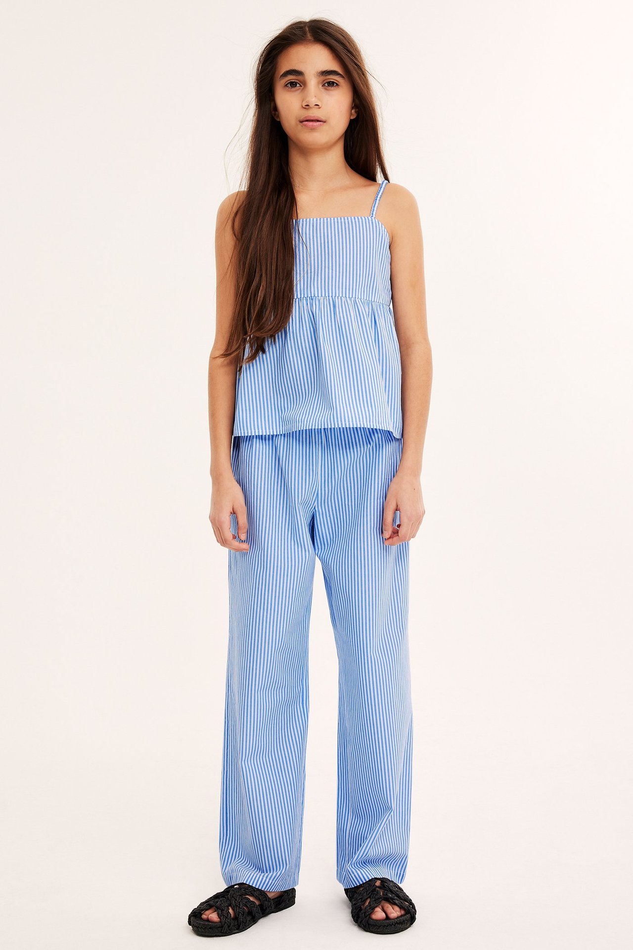 Framifrån: Ett barn i blåvitrandiga bomullsbyxor med resår, matchande linne och svarta flätade sandaler.