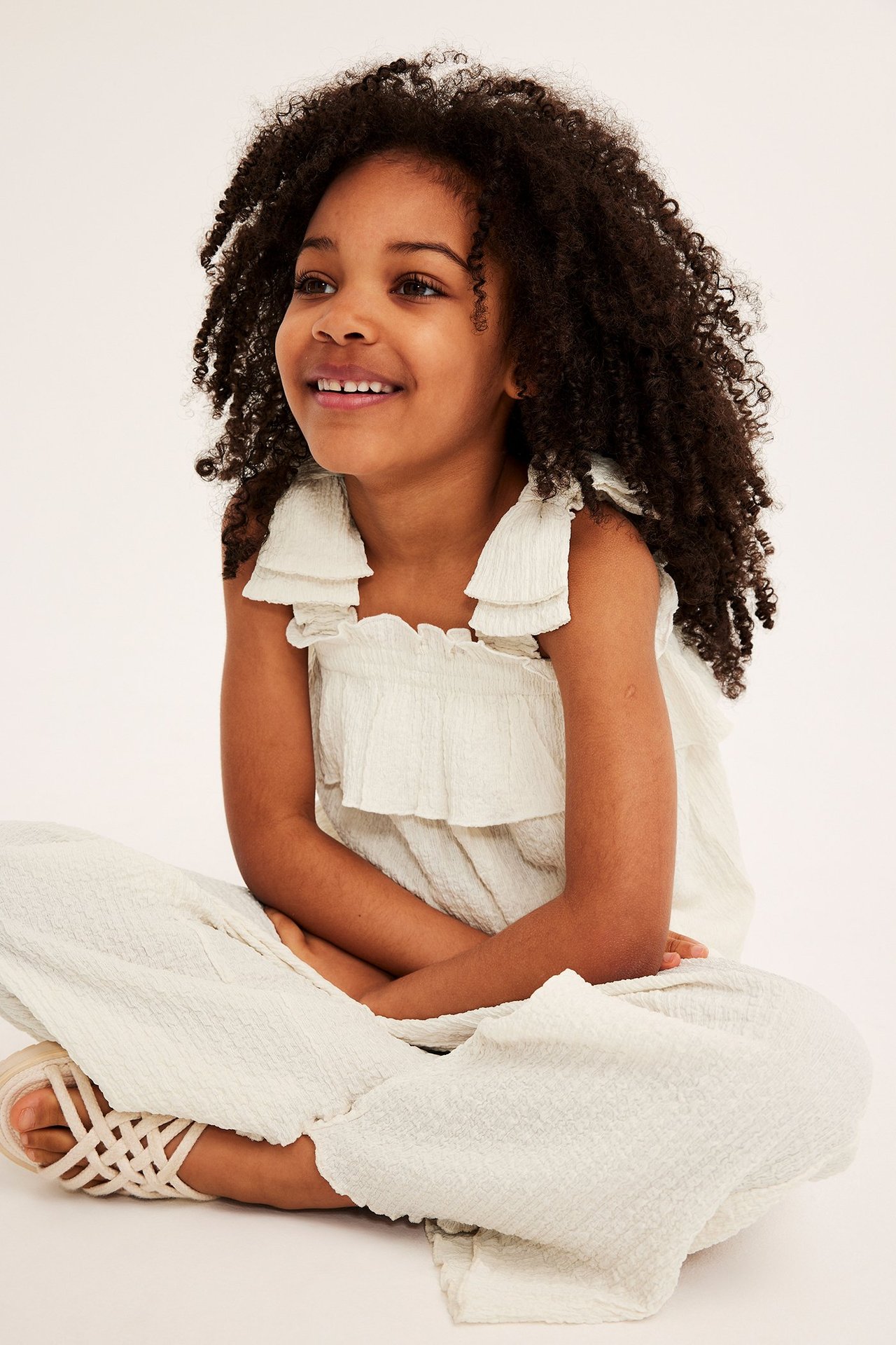 Widok z przodu: uśmiechnięte dziecko w ecru teksturowanym topie z falbankami, smockiem, pasujących spodniach i sandałach.