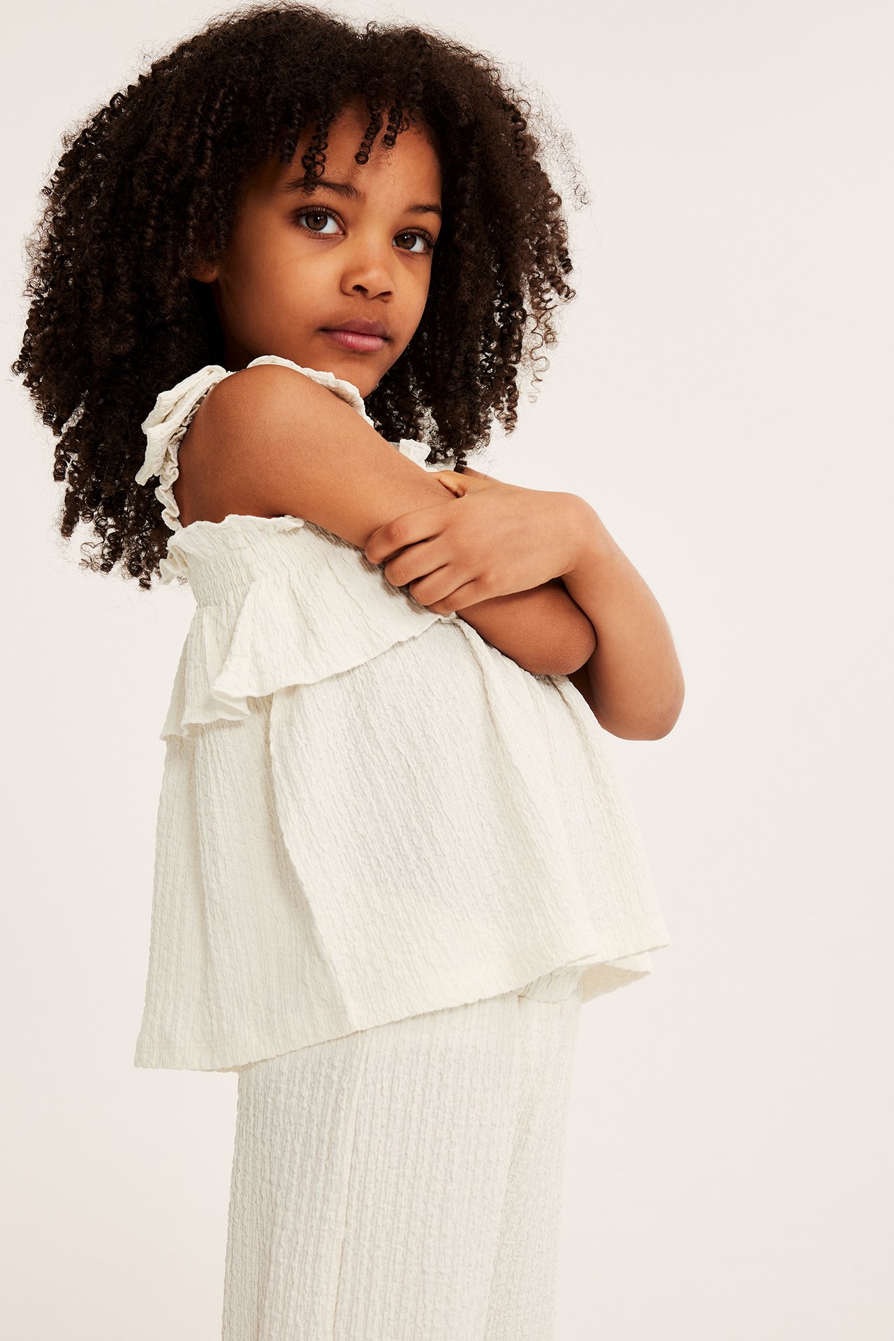 Sidovy av ett barn i offwhite texturerat linne med smock och volanger, samt matchande byxor.