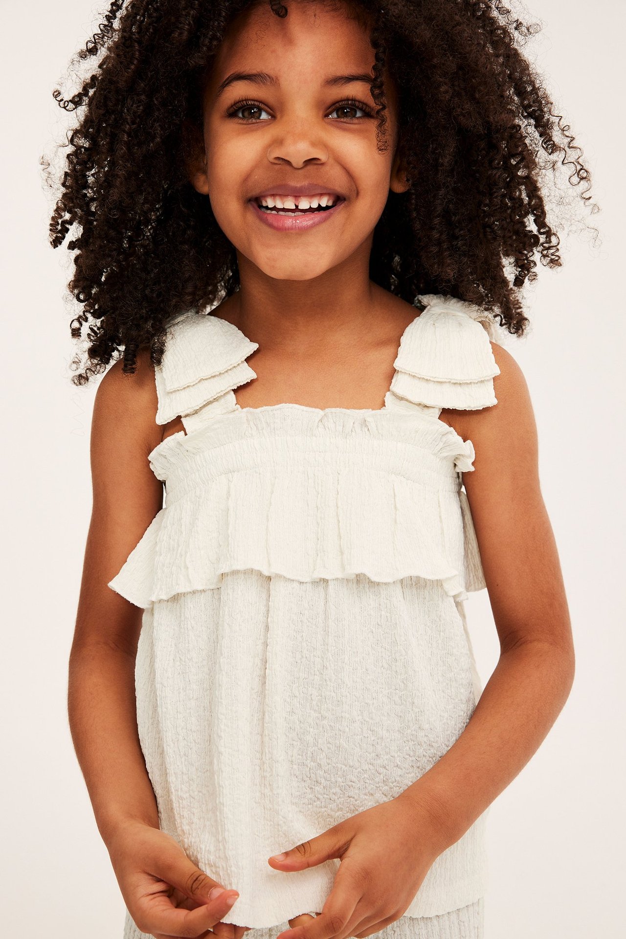 Widok z przodu dziecka w kremowym teksturowanym topie na ramiączkach z falbankami i marszczeniem.