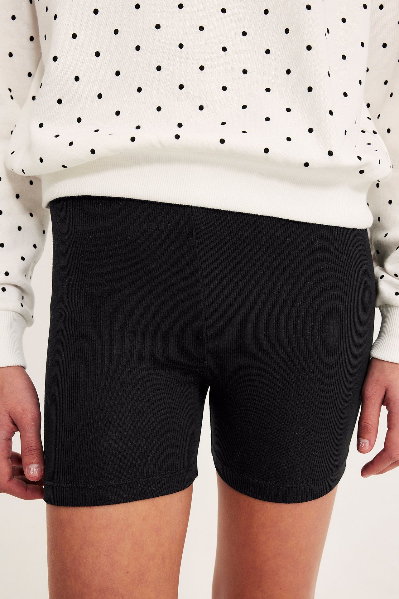 Forfra av et barn iført svarte ribbestrikkede sykkelshorts med en hvit prikkete genser.