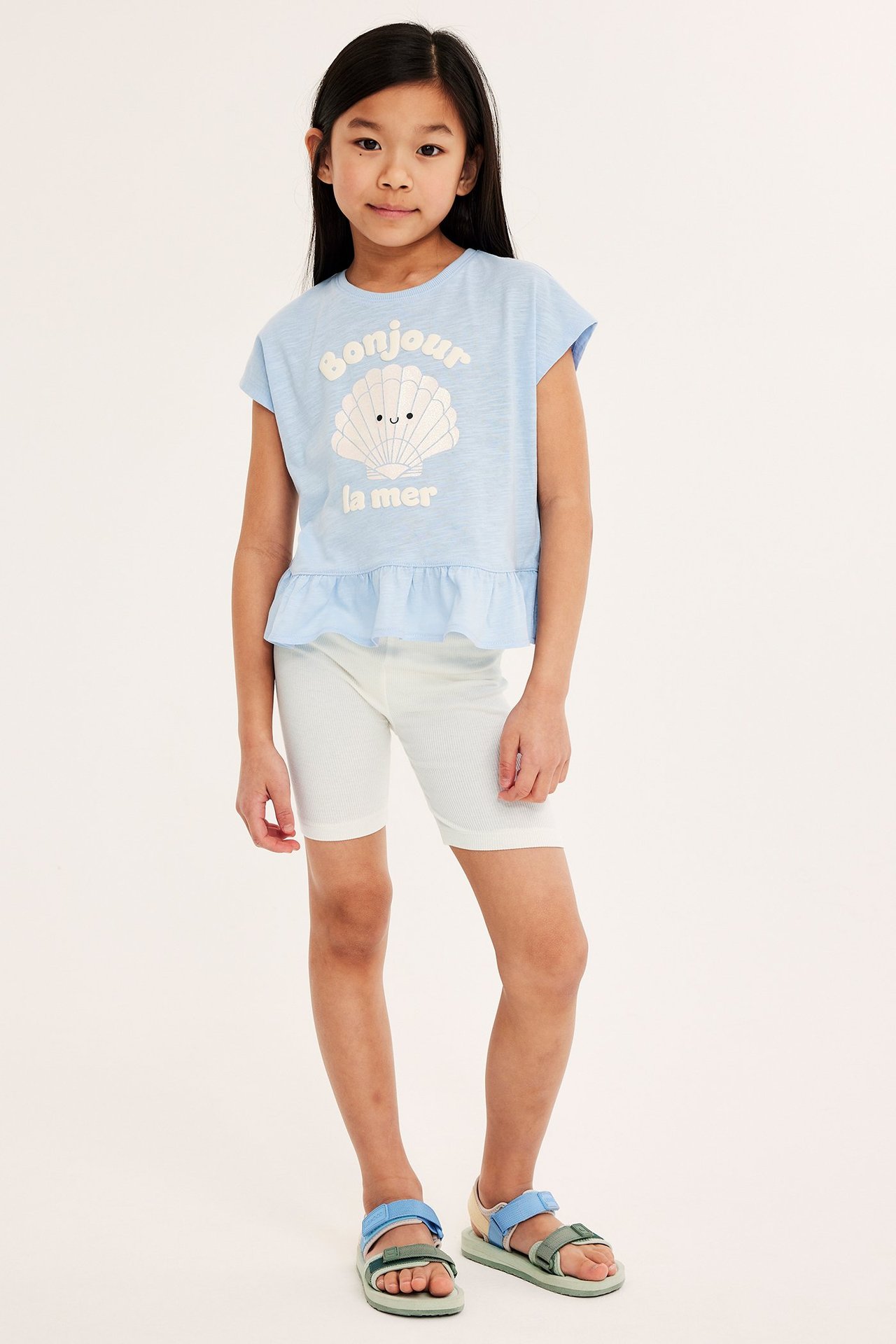 Edestä kuvattu lapsi yllään valkoiset ribatut pyöräilyshortsit, vaaleansininen simpukkakuvioinen t-paita ja moniväriset sandaalit.