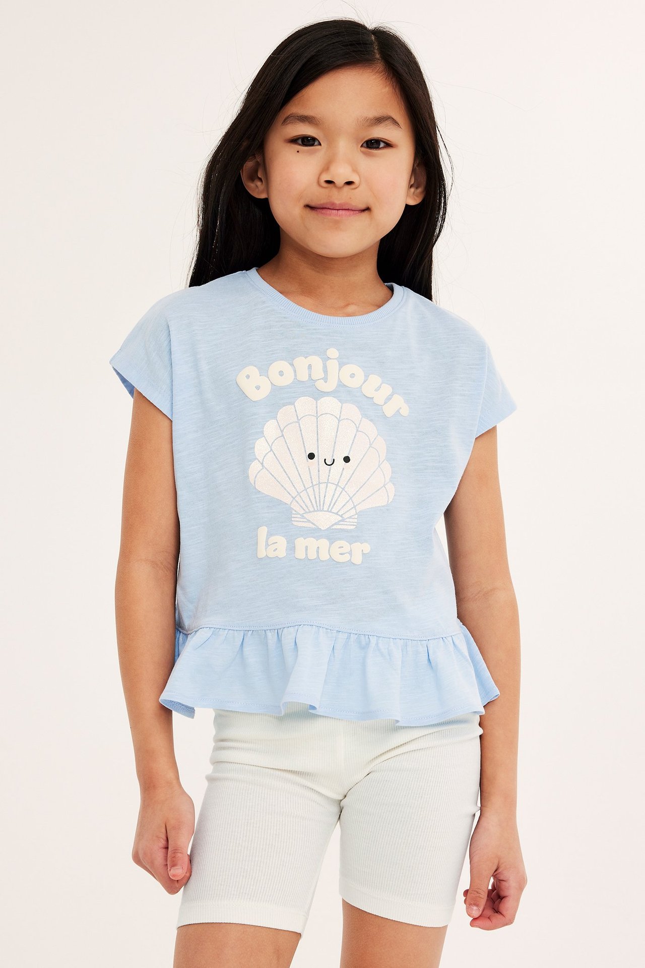 Edestä kuvattu lapsi valkoisissa ribatuissa pyöräilyshortseissa ja vaaleansinisessä t-paidassa simpukkakuviolla.