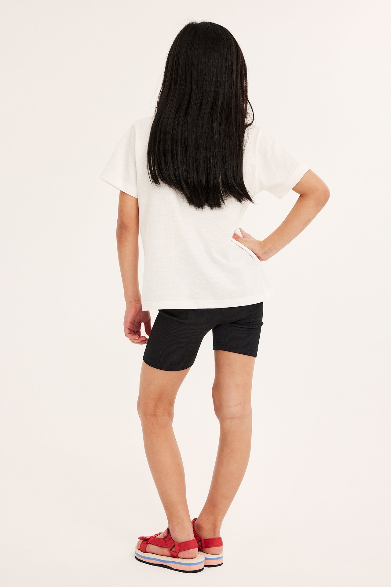 Bakfra av et barn i svarte ribbestrikkede sykkelshorts, en hvit t-skjorte og røde platåsandaler.
