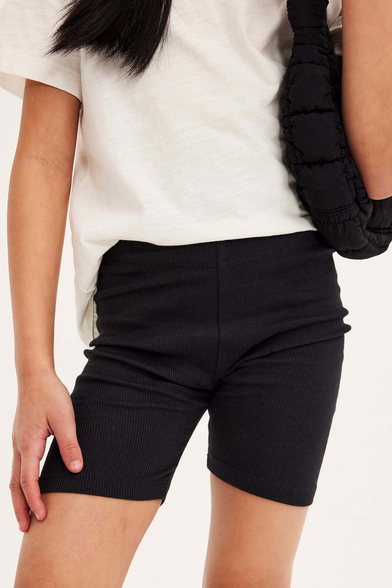 Edestä, lapsi yllään mustat ribbipyöräilyshortsit, valkoinen t-paita ja pitää mustaa topattua laukkua.