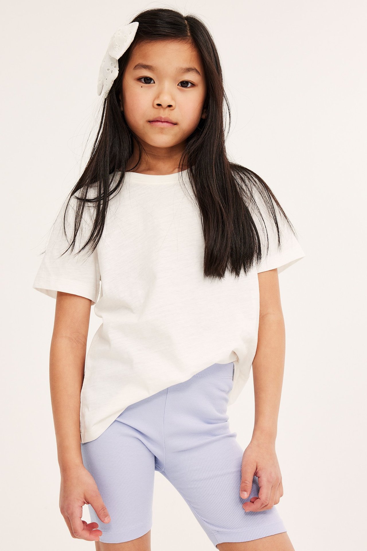Widok z przodu: Dziecko w liliowych prążkowanych kolarkach, białej koszulce i białej spince do włosów.