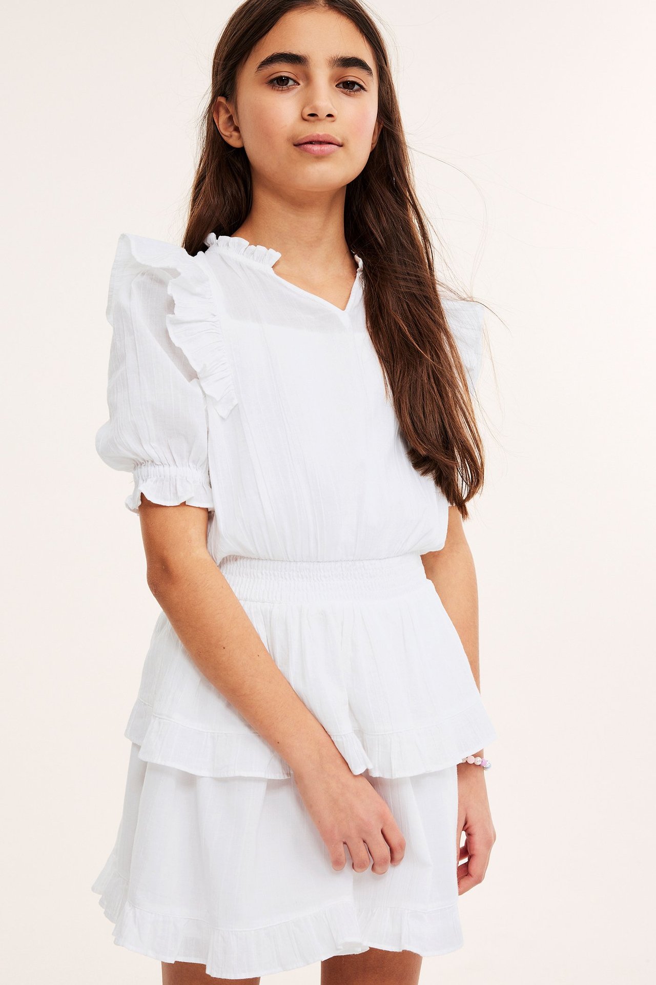 Forfra: et barn i hvit kortermet vevd kjole med volanger, strikk i midjen og et armbånd.