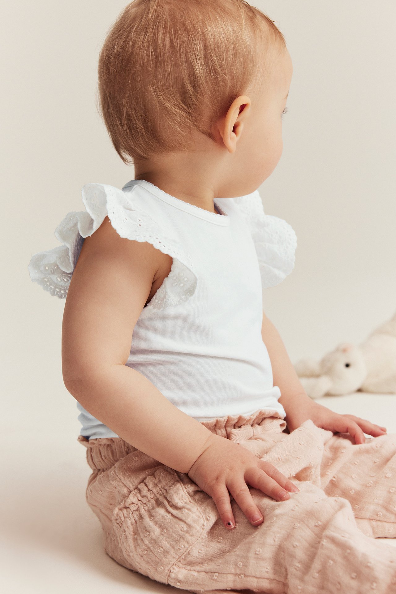 Sivukuva vauvasta luonnonvalkoisessa lyhythihaisessa bodyssä puuvillapitsisillä röyhelöhihoilla ja vaaleanpunaisissa kuvioituisissa housuissa.