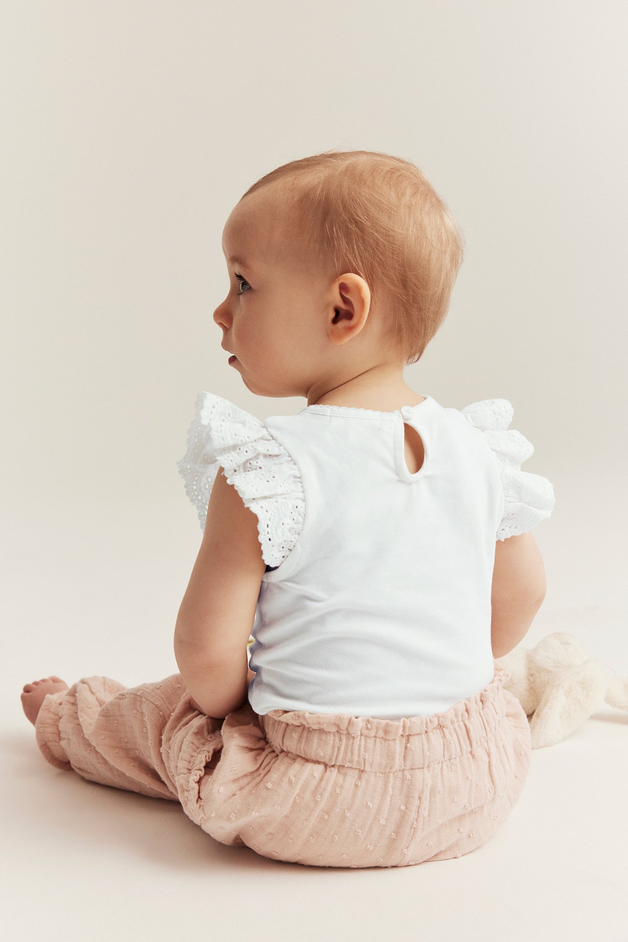 Takaa: vauva yllään luonnonvalkoinen lyhythihainen body pitsiröyhelöillä ja vaaleanpunaiset kuvioidut housut.