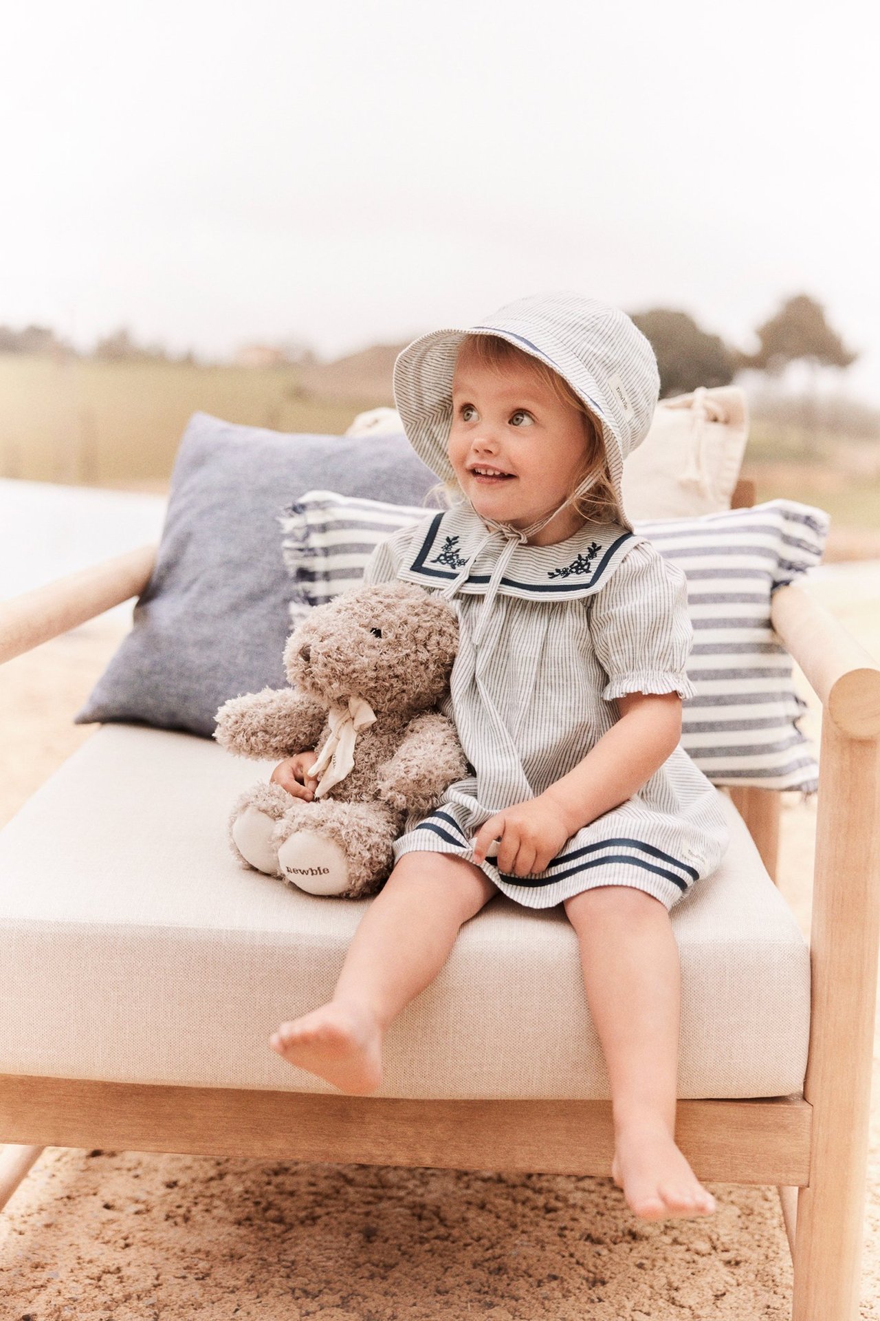 Forfra: Barn i offwhite stripet bomullskjole med sjømannskrage, matchende hatt, holder brun teddybjørn.