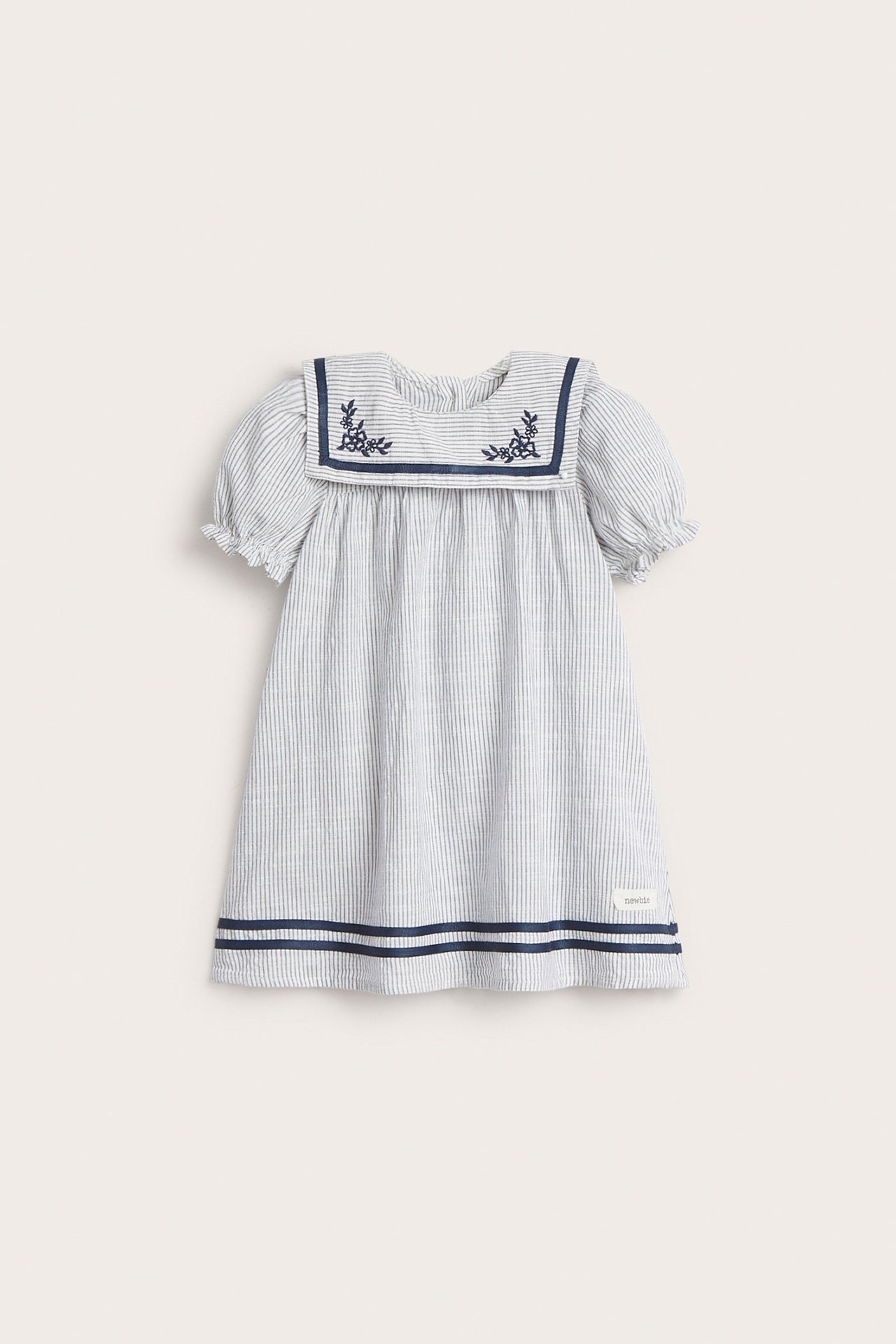 Frontvisning av en stripete bomullskjole for barn i offwhite med marineblå sjømannskrage, broderi, puffermer og kantbånd.