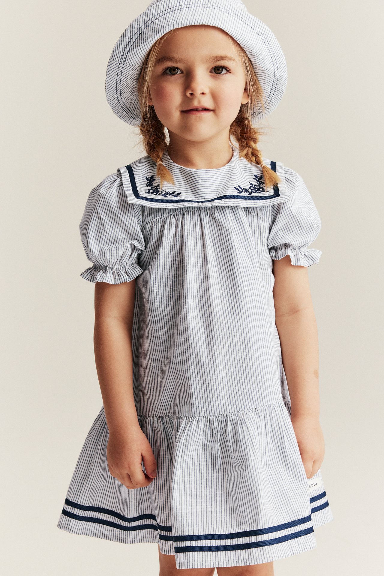 Framifrån: Barn i offwhite randig bomullsklänning med broderad sjömanskrage, puffärmar och matchande hatt.