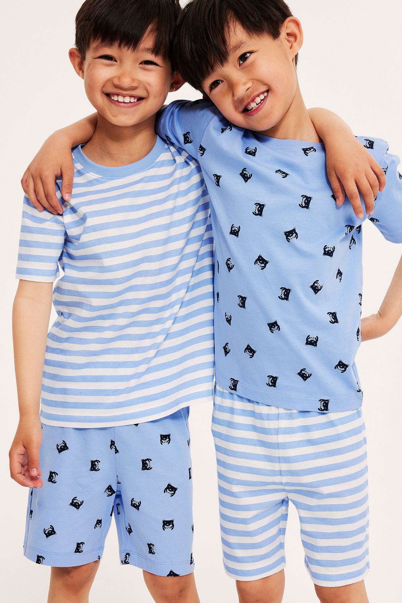 Edestä: kaksi hymyilevää lasta vaaleansinisissä puuvillapyjamissa, toinen raidallinen, toinen rapukuvioinen.