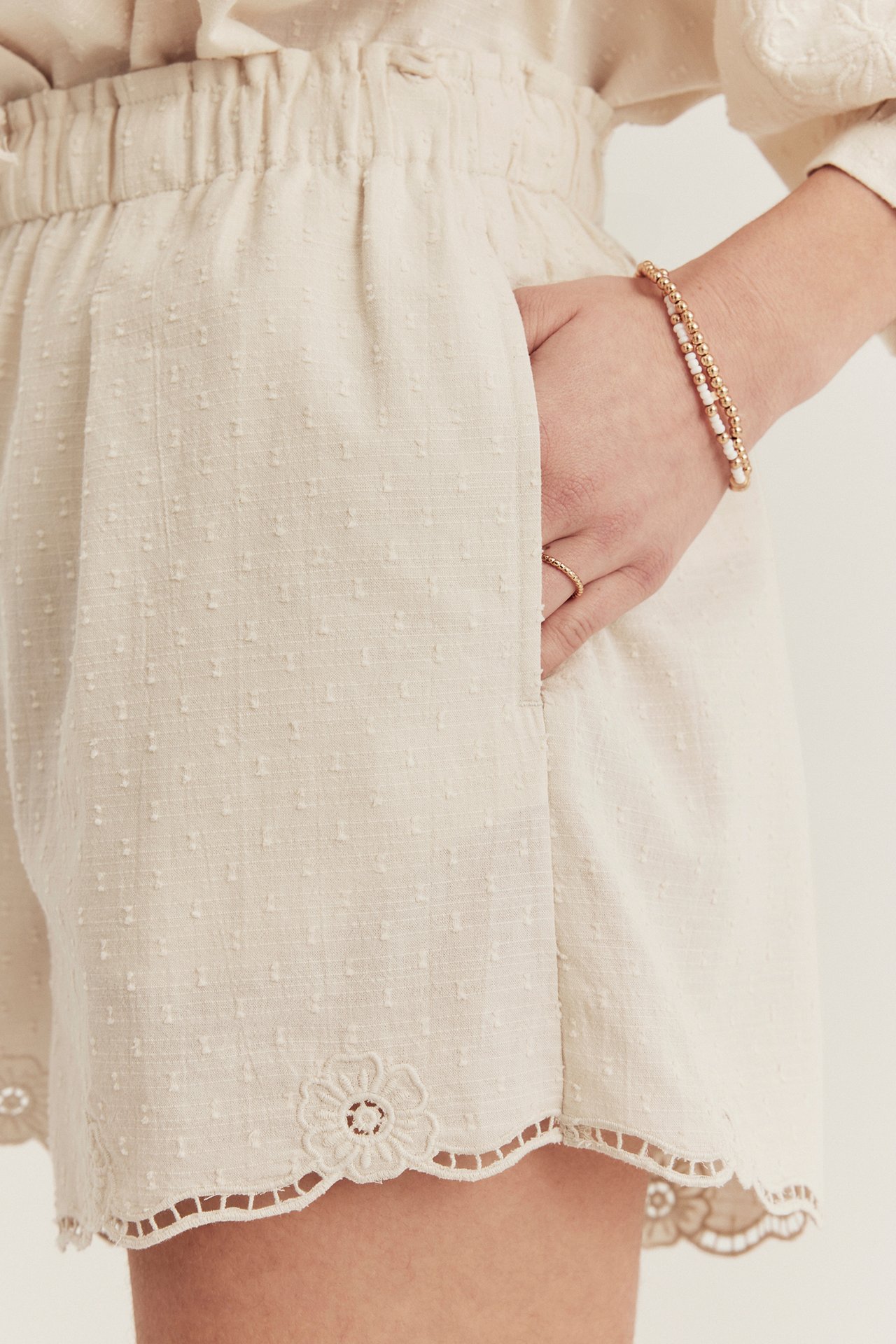 Närbild av kvinna i ljusbeige vävda shorts med prickig textur, resår i midjan, broderad fåll och pärlarmband.