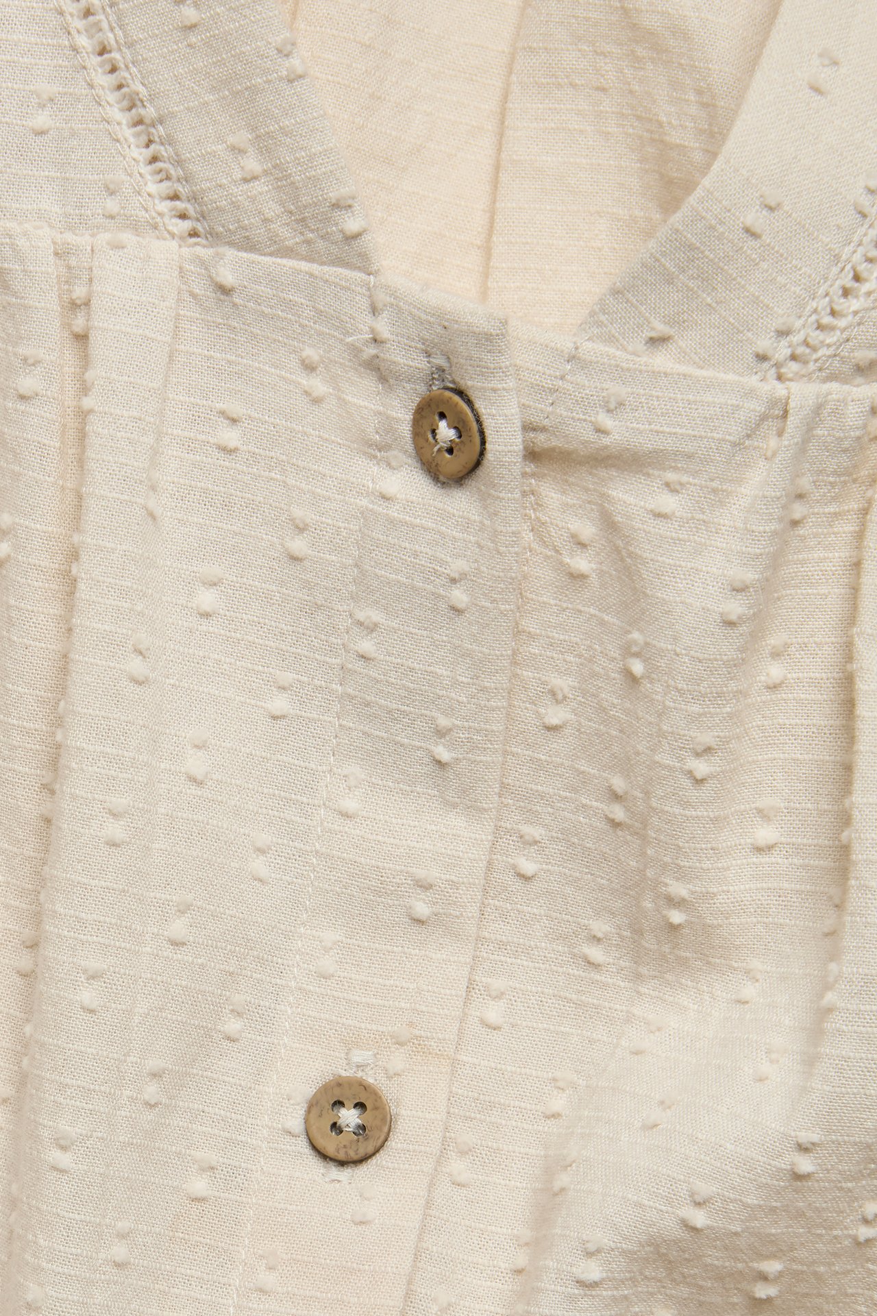 Närbild framifrån på en ljusbeige vävd blus med prickig textur, V-ringning med spets och knappar fram.